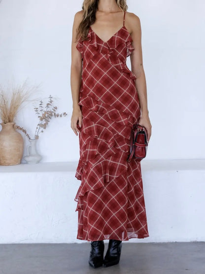 A woman models a rust-red maxi dress with a white plaid pattern and ruffled details, paired with black ankle boots and a matching small handbag, set against a white wall with decorative dried plants.