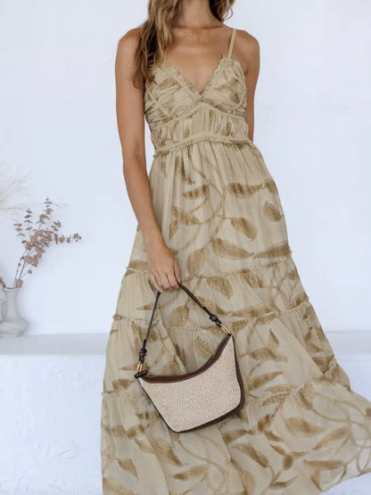 A woman wearing a beige maxi dress with a brown leaf pattern, holding a woven straw crescent handbag with dark brown trim, set against a bright white background.