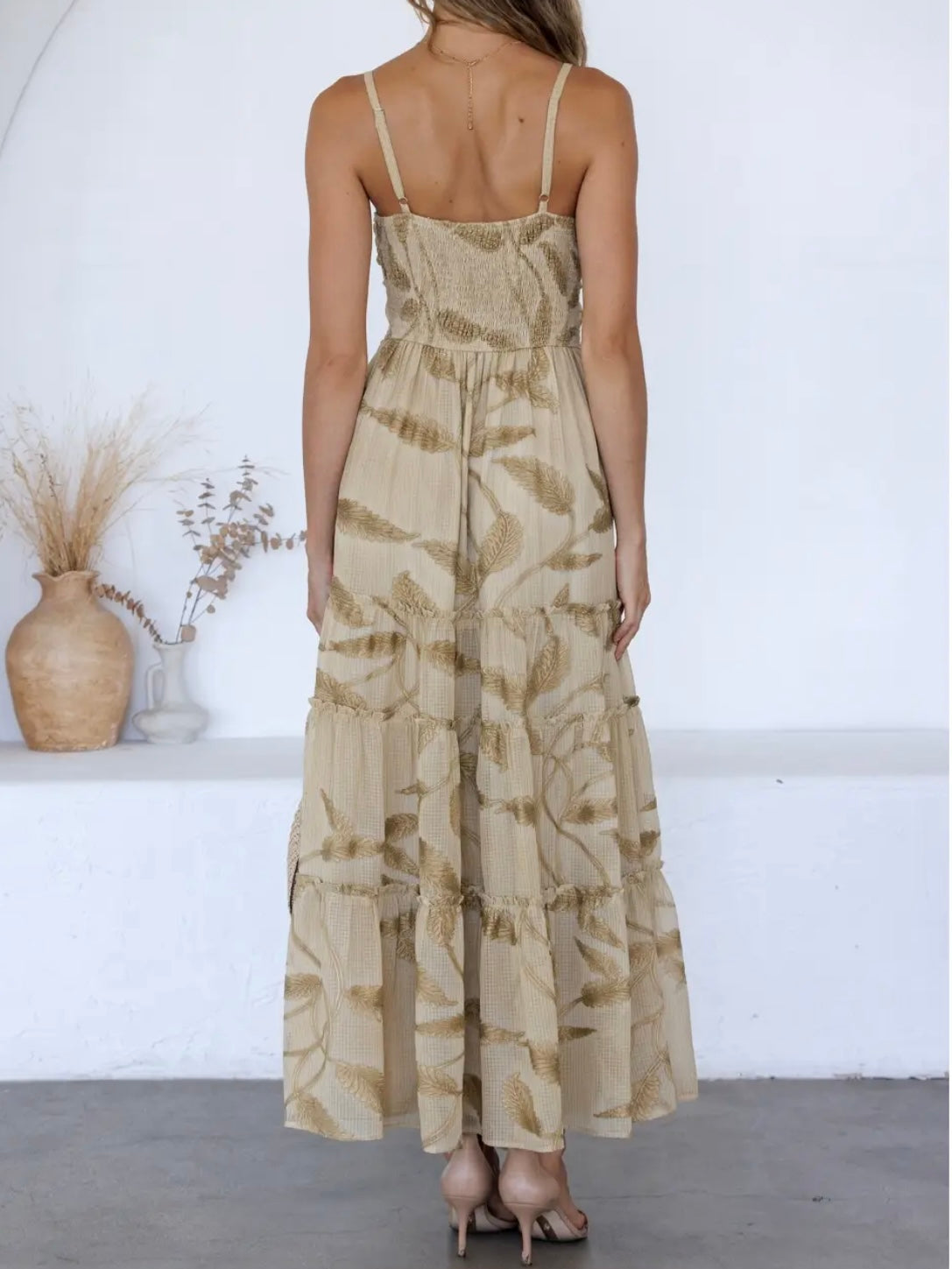 A woman shown from the back wearing a sleeveless, tiered maxi dress with a beige and brown leaf pattern, standing in a minimalist white room.