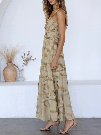 A woman models a sleeveless, tiered maxi dress with a beige and brown abstract pattern, paired with nude high-heeled sandals, set against a white wall with decorative vases.