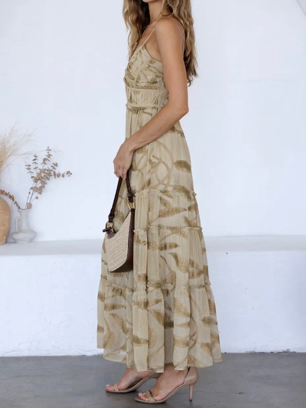 A woman wearing a beige and brown abstract patterned maxi dress, holding a brown and beige shoulder bag, standing in a bright white room.