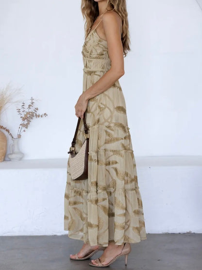 A woman wearing a beige and brown abstract patterned maxi dress, holding a brown and beige shoulder bag, standing in a bright white room.
