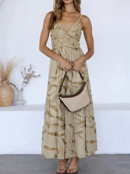 A woman wearing a beige maxi dress with a brown tie-dye pattern, holding a woven straw crescent handbag with a dark brown strap, standing against a white wall.