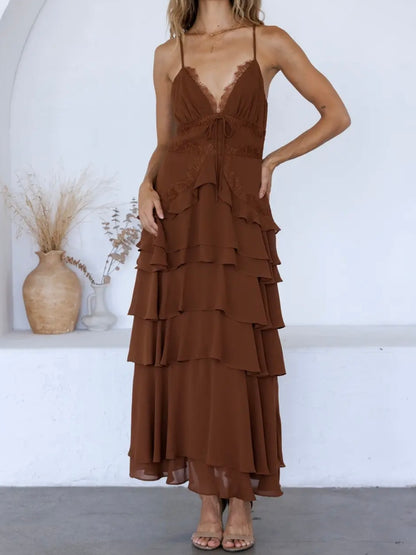 A woman models a brown tiered maxi dress with thin spaghetti straps and a lace V-neckline, standing in a bright white room with a vase of dried grass in the background.