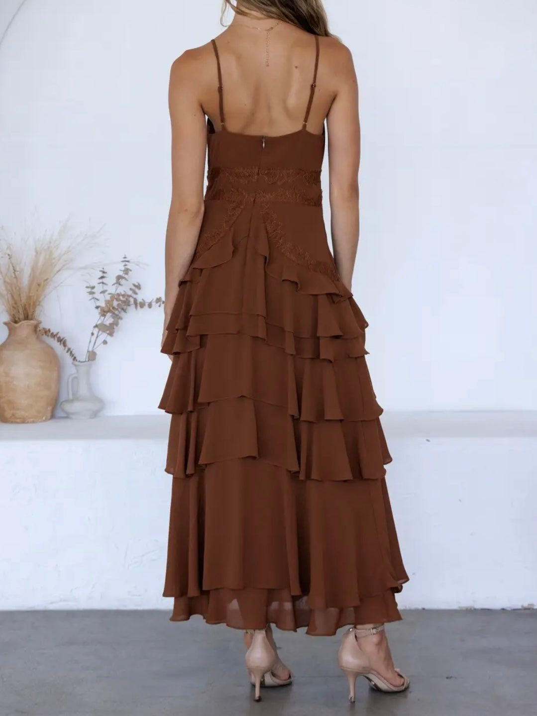 A woman shown from the back wearing a brown spaghetti-strap maxi dress with a ruffled tiered skirt, paired with beige high-heeled sandals, standing in a minimalist room with a white wall and floor.