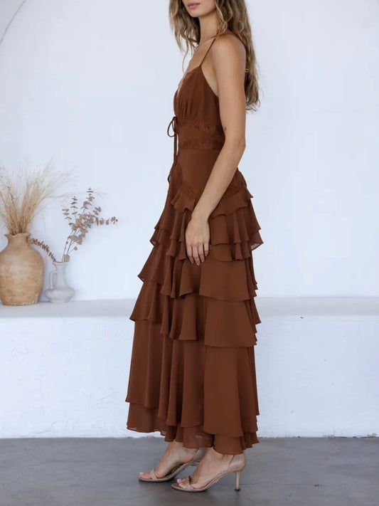 A woman wearing a brown sleeveless maxi dress with a tiered ruffle skirt, standing in a bright white room with decorative dried plants in a vase.