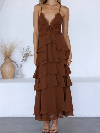 A woman wearing a brown tiered maxi dress with thin straps and a lace-trimmed V-neckline, standing against a white wall with decorative vases in the background.