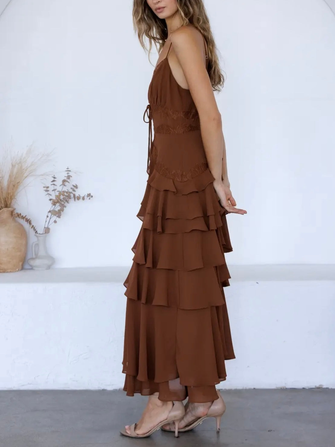 A woman wearing a brown tiered maxi dress with thin straps and a side tie, paired with nude high-heeled sandals, standing in a minimalist white room.