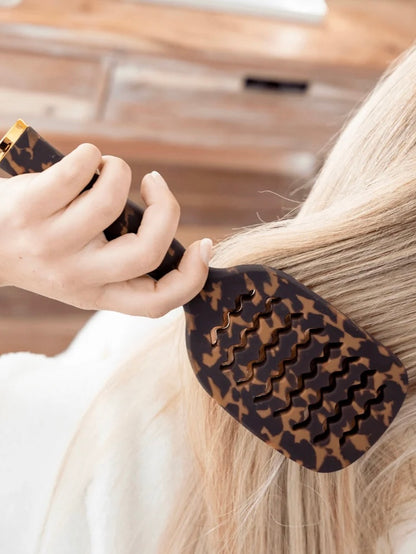 A close-up lifestyle shot of a person using a leopard-print hairbrush to brush their blonde hair. The hand holds the handle while the brush glides through the hair, with a softly blurred wooden background.