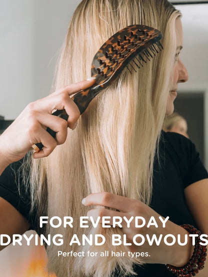 A woman with long blonde hair is shown from the back while being brushed with a wide-tooth hairbrush. The brush has a dark tortoiseshell pattern.