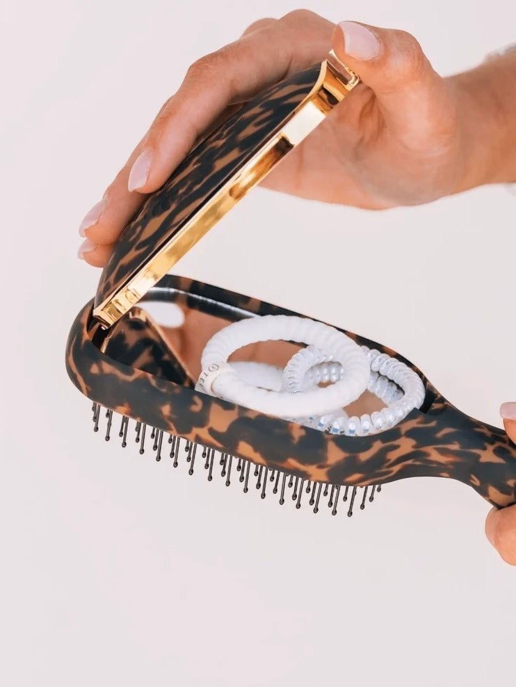 A close-up of a hand holding an open leopard-print hairbrush with gold-tone metal trim, showing white hair ties inside. The brush has a dark brown and tan leopard pattern with black bristles, set against a clean white background.