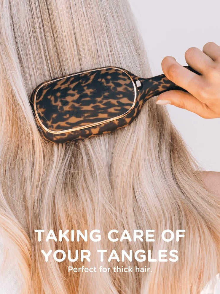 A close-up of a leopard-print hairbrush being used on long blonde hair, with a hand holding the handle. White text at the bottom reads 'TAKING CARE OF YOUR TANGLES' and 'Perfect for thick hair.
