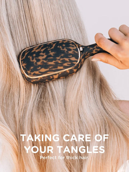 A close-up of a leopard-print hairbrush being used on long blonde hair, with a hand holding the handle. White text at the bottom reads 'TAKING CARE OF YOUR TANGLES' and 'Perfect for thick hair.