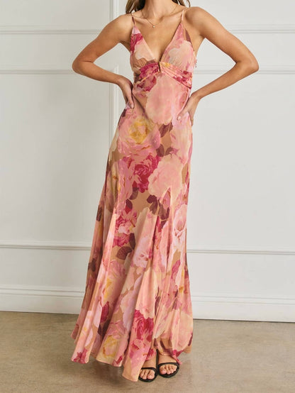 A woman modeling a sleeveless, floor-length maxi dress with a pink, peach, and red floral print, standing against a white paneled wall.