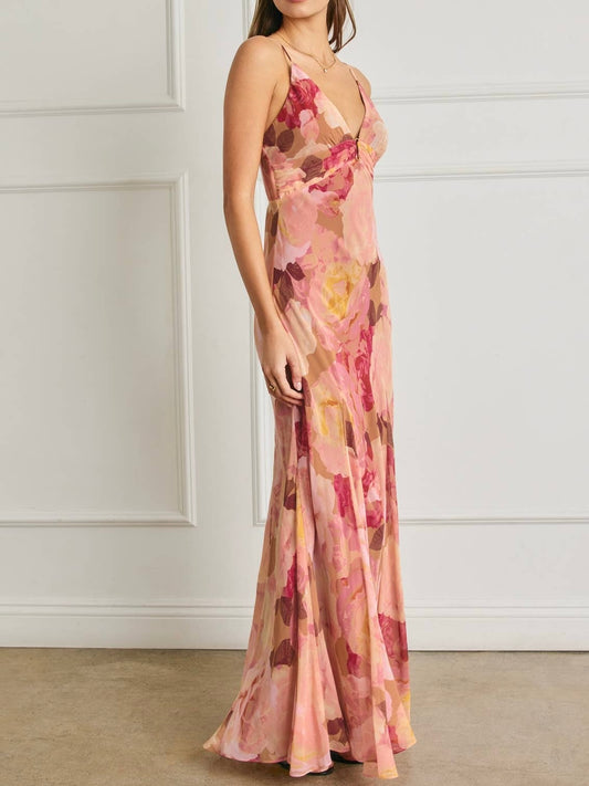 A woman modeling a sleeveless, floor-length pink floral gown with a V-neckline, set against a white paneled wall and beige floor.