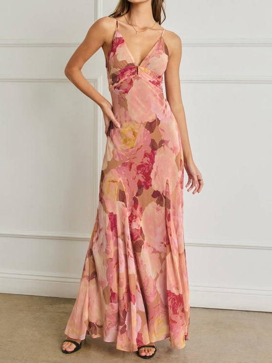 A woman modeling a sleeveless, floor-length maxi dress with a pink, peach, and red floral watercolor print, set against a white paneled wall.