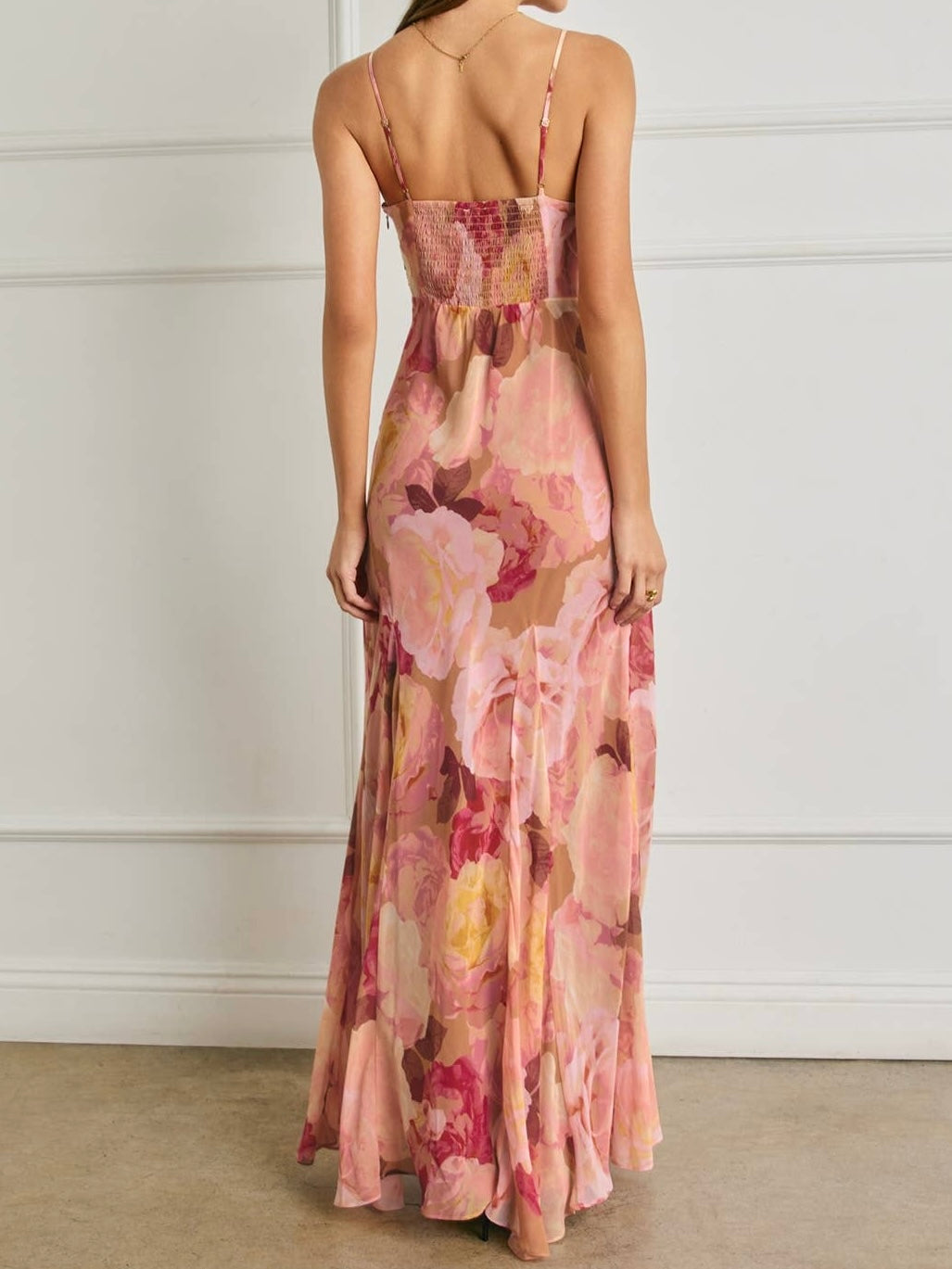 A woman modeling a long, backless maxi dress from the back, featuring a pink and peach floral watercolor print with thin spaghetti straps and a smocked bodice, set against a white paneled wall.